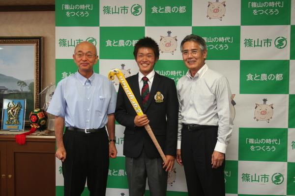 中央にホッケーのスティックを持った河野君、左側に市長、右側にYシャツ姿の男性が並んでいる写真